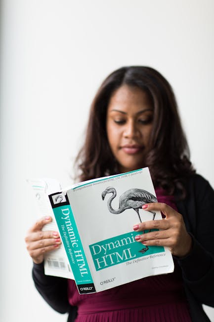 A woman with dark, shoulder-length hair is holding a book titled 'Dynamic HTML' by O'Reilly. She is dressed in a dark blazer over a maroon top and is looking at the book with a focused expression. The